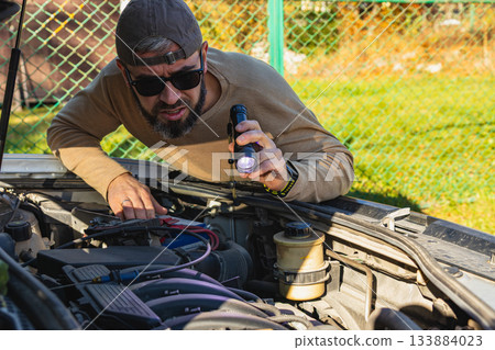 Man examines car engine in outdoor workshop with flashlight 133884023