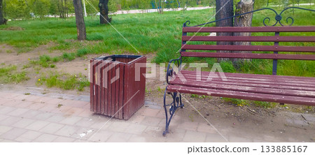 A rustic wooden bench and a slatted trash bin stand beside a tree on a paved path in a park. Green grass and trees form a natural, tranquil background 133885167