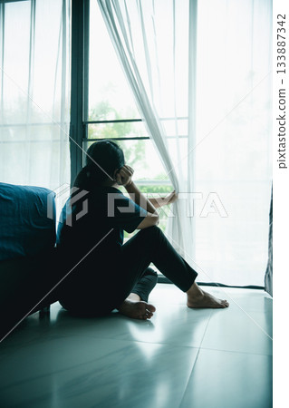 A lonely woman sits on the floor by window with hand on head in dark room. Concept of depression, sadness, anxiety, stress, mental health problem, loneliness and emotional struggle in life. 133887342