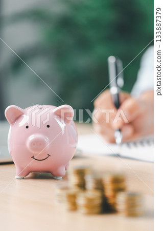 Concept of saving money and financial security. Pink piggy bank surrounded by coins on office desk with businesswoman in background, symbolizing investment, wealth growth and budget management. 133887379
