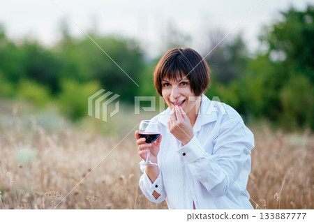 Brunette against the background of a dry field. Smiling girl with a glass of wine on vacation. Fruits, flowers and wine to lift your spirits.  133887777