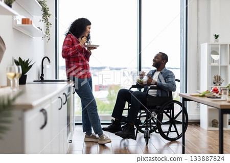 A diverse couple shares a meal in a bright, modern kitchen, with one person using a wheelchair. A diverse couple shares a meal in a bright, modern kitchen, with one person using a wheelchair. 133887824