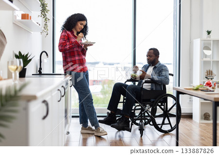 A diverse couple enjoys a healthy meal together in a modern kitchen setting, with one person using a wheelchair. 133887826