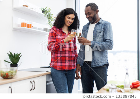 A smiling couple clinks glasses of wine in a modern kitchen, celebrating a special moment together. 133887843
