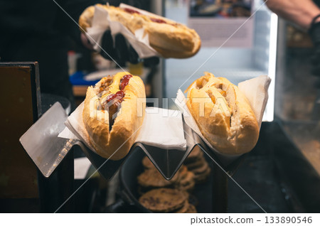 Street food scene with a chef making a fresh hot dog at a festive Christmas fair, surrounded by warm lights and holiday atmosphere. Street food scene with a chef making a fresh hot dog at a festive Christmas fair, surrounded by warm lights and holiday atmosphere. 133890546