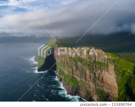 Scenic view of the Faroe Islands with dramatic green cliffs, rocky coastline, and ocean waves under cloudy northern weather. Rugged, untouched nature of the Nordladalur 133890764