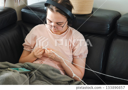 Teenager girl wearing glasses and headphones sitting comfortably at home, knitting using a colorful yarn, following a pattern or scheme displayed on her smartphone. Cozy creative hobby scene. Teenager girl wearing glasses and headphones sitting comfortably at home, knitting using a colorful yarn, following a pattern or scheme displayed on her smartphone. Cozy creative hobby scene. 133890857