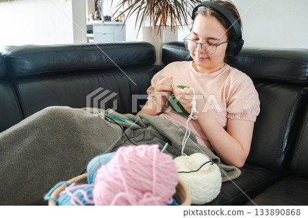 Teenager girl wearing glasses and headphones sitting comfortably at home, knitting using a colorful yarn, following a pattern or scheme displayed on her smartphone. Cozy creative hobby scene. 133890868