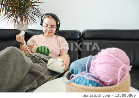 Teenager girl wearing glasses and headphones sitting comfortably at home, knitting using a colorful yarn, following a pattern or scheme displayed on her smartphone. Cozy creative hobby scene. 133890869