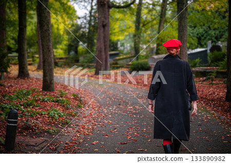 Elegant, well-groomed woman with fashionable style walking in an autumn park on a cloudy day. She wears a red beret, red lipstick, and matching red tights, looking confident and sophisticated. Elegant, well-groomed woman with fashionable style walking in an autumn park on a cloudy day. She wears a red beret, red lipstick, and matching red tights, looking confident and sophisticated. 133890982