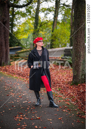 Elegant, well-groomed woman with fashionable style walking in an autumn park on a cloudy day. She wears a red beret, red lipstick, and matching red tights, looking confident and sophisticated. Elegant, well-groomed woman with fashionable style walking in an autumn park on a cloudy day. She wears a red beret, red lipstick, and matching red tights, looking confident and sophisticated. 133891007