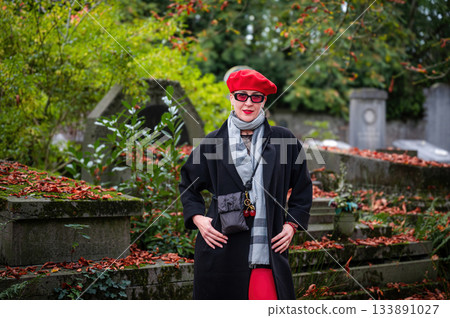 Elegant, well-groomed woman with fashionable style walking in an autumn park on a cloudy day. She wears a red beret, red lipstick, and matching red tights, looking confident and sophisticated. 133891027
