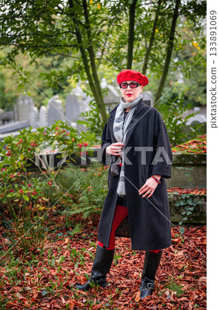 Elegant, well-groomed woman with fashionable style walking in an autumn park on a cloudy day. She wears a red beret, red lipstick, and matching red tights, looking confident and sophisticated. 133891069