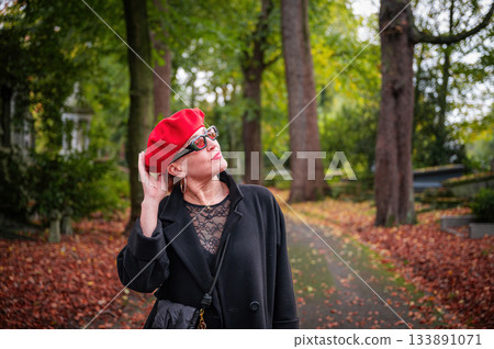 Elegant, well-groomed woman with fashionable style walking in an autumn park on a cloudy day. She wears a red beret, red lipstick, and matching red tights, looking confident and sophisticated. Elegant, well-groomed woman with fashionable style walking in an autumn park on a cloudy day. She wears a red beret, red lipstick, and matching red tights, looking confident and sophisticated. 133891071