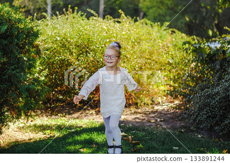 Adorable small girl wearing a white dress and diadem, happily playing outdoors in a sunny green park. Adorable small girl wearing a white dress and diadem, happily playing outdoors in a sunny green park. 133891244
