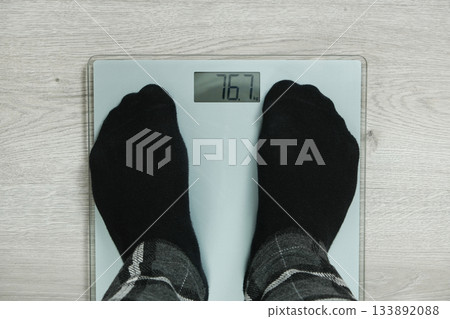 Top View of Feet in Black Socks Standing on Digital Bathroom Scales, Showing Weight 76.7 KG. 133892088