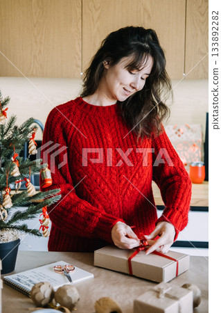 Young adult woman smiles while carefully tying ribbon on wrapped gift box on kitchen counter next to decorated tree. Packaging orders, Christmas side hustle, small brand aesthetic, festive gifting. Young adult woman smiles while carefully tying ribbon on wrapped gift box on kitchen counter next to decorated tree. Packaging orders, Christmas side hustle, small brand aesthetic, festive gifting. 133892282