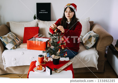 A young woman in a Santa hat and red sweater sits on a sofa decorating a small tabletop Christmas tree. Festive decor, small space living, generational traditions, seasonal homemaking. 133892285