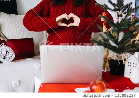 Young woman in red sweater forming a heart shape with her hands in front of a laptop during the Christmas season. Digital connection, festive self-expression, online celebrations, cozy holiday mood 133892287