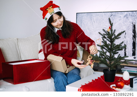 A young woman sits on a sofa decorating a small tabletop Christmas tree with miniature ornaments. Mindful decorating, intentional holiday prep, slow living, thoughtful holiday rituals. A young woman sits on a sofa decorating a small tabletop Christmas tree with miniature ornaments. Mindful decorating, intentional holiday prep, slow living, thoughtful holiday rituals. 133892291