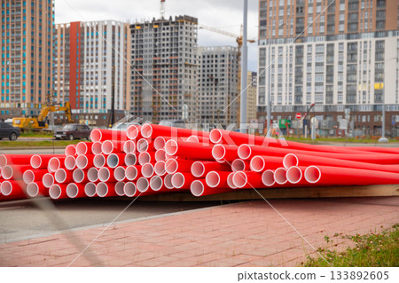 Stack of red new plastic pipes on street. Modern buildings rise in background, urban development, street repair. 133892605