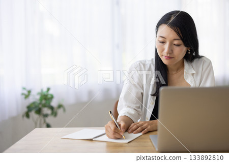 一位女士一邊在電腦上工作，一邊在筆記本上做筆記。 133892810