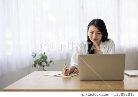 一位女士一邊在電腦上工作，一邊在筆記本上做筆記。 133892812