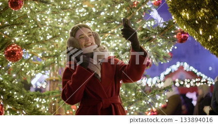 A young beautiful girl in a red coat walks in the evening at the European Christmas Market making a video call standing against the backdrop of a Christmas tree with garlands and illuminations A young beautiful girl in a red coat walks in the evening at the European Christmas Market making a video call standing against the backdrop of a Christmas tree with garlands and illuminations 133894016