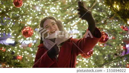 A young beautiful girl in a red coat walks in the evening at the European Christmas Market making a video call standing against the backdrop of a Christmas tree with garlands and illuminations 133894022