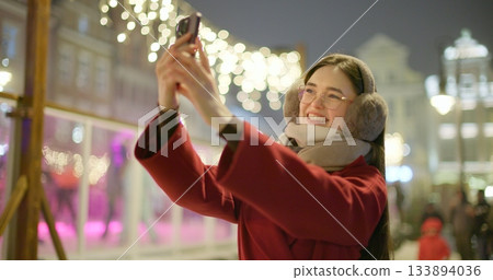 A young beautiful girl in a red coat walks in the evening at the European Christmas Market and talks on the phone against the backdrop of an ice rink with illumination and garlands. 133894036