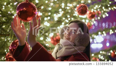 A young beautiful girl in a red coat stands near a Christmas tree with garlands and illuminations in the evening at the European Christmas Market and touches the Christmas tree decorations. 133894052