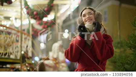 A young beautiful girl in a red coat walks in the evening at the European Christmas Market against the backdrop of a festive carousel with illuminations. A young beautiful girl in a red coat walks in the evening at the European Christmas Market against the backdrop of a festive carousel with illuminations. 133894056