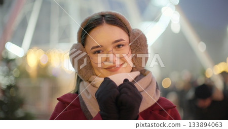 A young beautiful girl in a red coat walks in the evening at the New Year's Christmas fair against the backdrop of a Ferris wheel among lights and garlands and admires the decorations 133894063