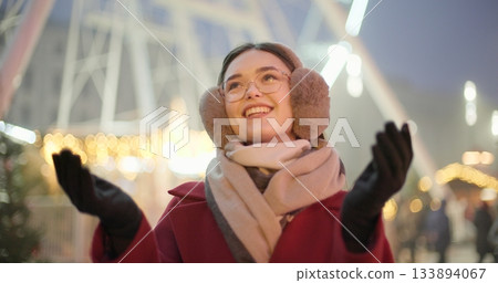A young beautiful girl in a red coat walks in the evening at the New Year's Christmas fair against the backdrop of a Ferris wheel among lights and garlands and admires the decorations A young beautiful girl in a red coat walks in the evening at the New Year's Christmas fair against the backdrop of a Ferris wheel among lights and garlands and admires the decorations 133894067
