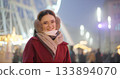 A young beautiful girl in a red coat walks in the evening at the New Year's Christmas fair against the backdrop of a Ferris wheel among lights and garlands and admires the decorations 133894070