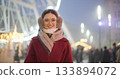 A young beautiful girl in a red coat walks in the evening at the New Year's Christmas fair against the backdrop of a Ferris wheel among lights and garlands and admires the decorations 133894072