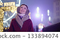 A young beautiful girl in a red coat walks in the evening at the New Year's Christmas fair against the backdrop of a Ferris wheel among lights and garlands and admires the decorations 133894074