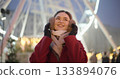 A young beautiful girl in a red coat walks in the evening at the New Year's Christmas fair against the backdrop of a Ferris wheel among lights and garlands and admires the decorations 133894076