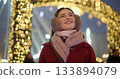 A young beautiful girl in a red coat walks in the evening at the New Year's Christmas fair against the backdrop of a Ferris wheel among lights and garlands and admires the decorations 133894079