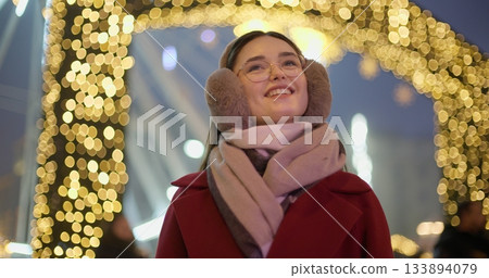 A young beautiful girl in a red coat walks in the evening at the New Year's Christmas fair against the backdrop of a Ferris wheel among lights and garlands and admires the decorations 133894079