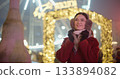 A young beautiful girl in a red coat walks in the evening at the New Year's Christmas fair against the backdrop of a Ferris wheel among lights and garlands and admires the decorations 133894082