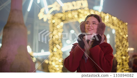 A young beautiful girl in a red coat walks in the evening at the New Year's Christmas fair against the backdrop of a Ferris wheel among lights and garlands and admires the decorations A young beautiful girl in a red coat walks in the evening at the New Year's Christmas fair against the backdrop of a Ferris wheel among lights and garlands and admires the decorations 133894082
