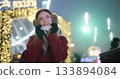 A young beautiful girl in a red coat walks in the evening at the New Year's Christmas fair against the backdrop of a Ferris wheel among lights and garlands and admires the decorations 133894084