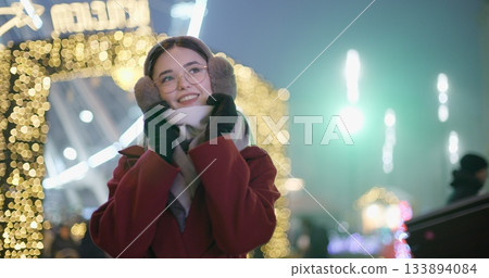 A young beautiful girl in a red coat walks in the evening at the New Year's Christmas fair against the backdrop of a Ferris wheel among lights and garlands and admires the decorations A young beautiful girl in a red coat walks in the evening at the New Year's Christmas fair against the backdrop of a Ferris wheel among lights and garlands and admires the decorations 133894084