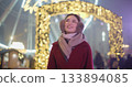 A young beautiful girl in a red coat walks in the evening at the New Year's Christmas fair against the backdrop of a Ferris wheel among lights and garlands and admires the decorations 133894085
