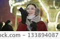 A young beautiful girl in a red coat walks in the evening at the Christmas fair and talks on the phone or records a voice message standing against the backdrop of a carousel with illuminations 133894087