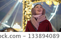 A young beautiful girl in a red coat walks in the evening at the New Year's Christmas fair against the backdrop of a Ferris wheel among lights and garlands and admires the decorations 133894089