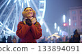 A young beautiful girl in a red coat walks in the evening at the New Year's Christmas fair against the backdrop of a Ferris wheel among lights and garlands and admires the decorations 133894091
