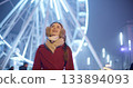 A young beautiful girl in a red coat walks in the evening at the New Year's Christmas fair against the backdrop of a Ferris wheel among lights and garlands and admires the decorations 133894093