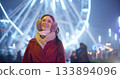 A young beautiful girl in a red coat walks in the evening at the New Year's Christmas fair against the backdrop of a Ferris wheel among lights and garlands and admires the decorations 133894096